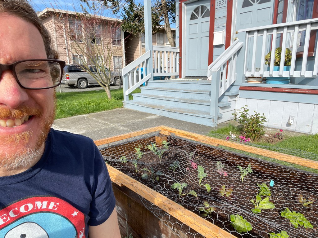 Color photo of smiling, bearded man on the left edge of the frame in front of newly planted garden with framed chicken wire to keep cats from soiling the soil in the front yard of a light blue house with red trim and quaint (or neglected) worn steps leading up to two doors of the three units contained therein, known in some circles as the Blue Raspberry House.